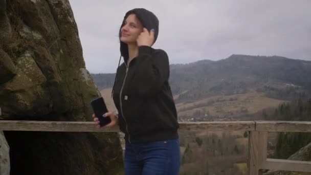 Mujer joven adulta de pie en las montañas y sonriendo mirando a la cámara. Turista disfrutando del paisaje de montaña sosteniendo el teléfono móvil en las manos. — Vídeos de Stock