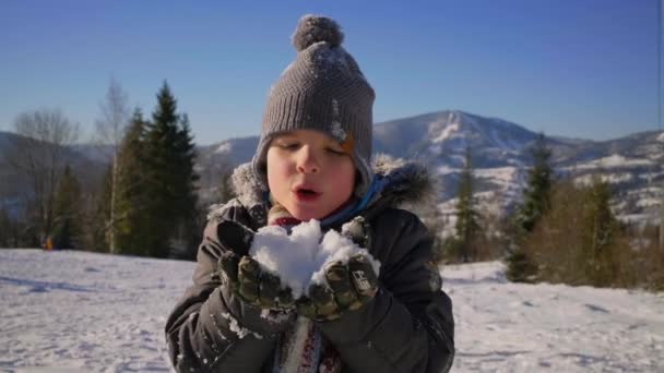 Портрет улыбающегося ребенка, дующего на снегу на зимней горе в солнечный холодный день. Мальчик брызгает снегом. Зимние семейные мероприятия. — стоковое видео