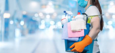 Cleaning lady in a protective mask with a bucket and cleaning products on a blurred background.