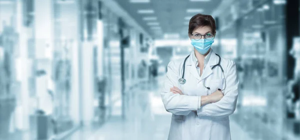 A doctor in a mask with his arms crossed stands on a blurred background of the hospital.