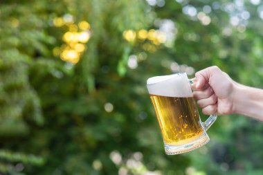 A glass of beer in a hand on a blurred background of trees.