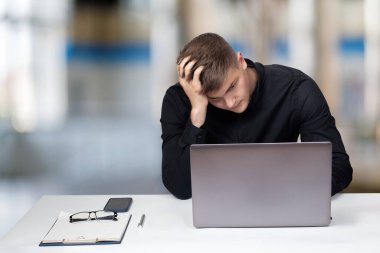 Concept of crisis in business at work. Young businessman holding his head sitting at a table in front of a computer.