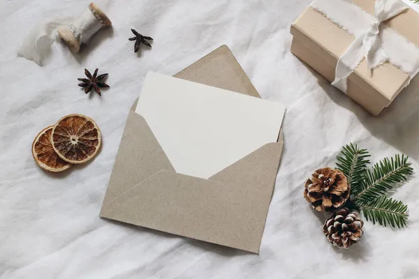 Kerstcadeau inpakken stilleven. Spar boom takken, aanwezig doos en droge sinaasappelschijfjes. Blanco kaart model, ambachtelijke enveloppe en dennenappels. Witte linnen tafelkleed achtergrond. Flatlay, bovenaanzicht. — Stockfoto