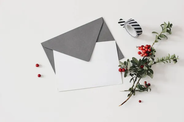 Λευκά ευχετήρια κάρτα mockup με craft φάκελο και washi ταινία. Χριστουγεννιάτικα χαρτικά, λίστα επιθυμιών. Κόκκινα φρούτα lingonberries με πράσινα φύλλα που απομονώνονται σε λευκό φόντο πίνακα. Χειμερινό επίπεδο lay, κορυφαία άποψη — Φωτογραφία Αρχείου
