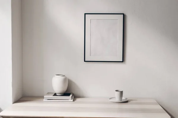 Elegáns minimális otthoni irodai csendélet kompozíció. Fehér váza, egy csésze kávé, magazinok. Fa asztal háttér napfényben. Üres fekete képkeret, fehér nappali fal. Skandináv belső tér. — Stock Fotó