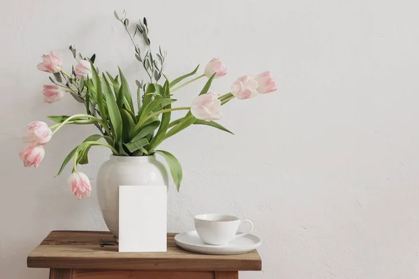 イースター、春の静物画。ヴィンテージ木製のベンチに白いセラミック花瓶にコーヒーと花の花束のカップ。空のグリーティングカードモックアップ。ピンクチューリップ、テーブルの上のオリーブの木の枝. — ストック写真