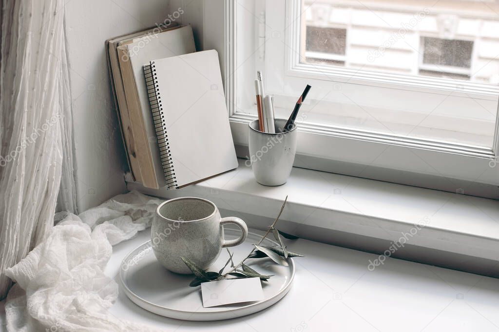 Home office still life. Blank diary and business card mockup. Cup of coffee, olive tree branch on tray. Books and pencils in ceramic mug on window sill. Scandinavian interior.. Cotton muslin curtain.