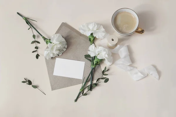 Breakfast still life. Greeting card, invitation mockup with envelope. Wedding, birthday stationery. Cup of coffee, white carnation flowers and eucalyptus branches. Beige table background. Flat lay. — Stock Photo, Image
