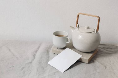 Kahvaltı natürmort. Bir fincan kahve, seramik çaydanlığı. Boş tebrik kartı, davetiye maketi. Bej keten masa örtüsü. Beyaz duvar arka planda. Tarafsız sabah yaşam tarzı kompozisyonu.