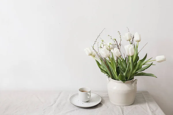 Páscoa feminina, primavera ainda cena da vida. Xícara de café e buquê floral. vaso de cerâmica branca na mesa. Flores de tulipas brancas, ramos de cerejeira em toalha de mesa de linho. Interior com estilo. Espaço de cópia. — Fotografia de Stock