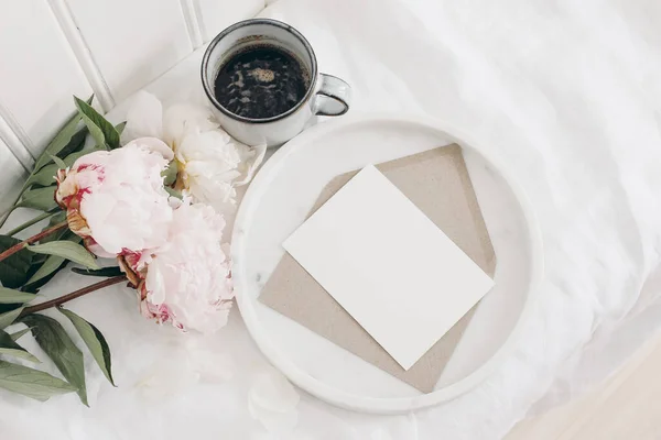 Kvetoucí růžové pivoňky, kytice ležící na stole. Bílá dřevěná zeď. Prázdný pozdrav na mramorovém podnosu. Šálek kávy, snídaně do postele. Svatební nebo narozeninová oslava, koncept dárků. — Stock fotografie