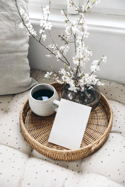 Spring still life composition. Greeting card mockup on wickered tray, cup of coffee. Feminine styled photo. Floral scene with blurred white cherry tree blossoms on bench near window. Selective focus — Stock Photo, Image