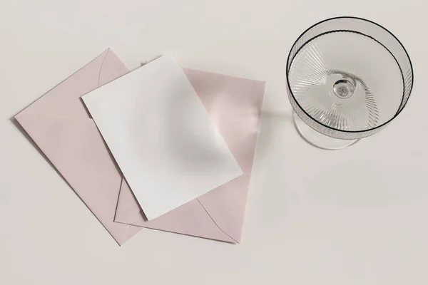 Σύγχρονη καλοκαιρινή γραφική ύλη νεκρή φύση. Σαμπάνια, ποτήρι με μακριές σκιές. Ροζ φάκελοι, λευκή ευχετήρια κάρτα, μακιγιαρισμένη σκηνή. Μπεζ φόντο πίνακα στο φως του ήλιου. Επίπεδο lay, πάνω όψη. — Φωτογραφία Αρχείου