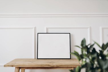 Blank horizontal black picture frame mockup. Old wooden console table. White wall moulding background, trim decor. Elegant home interior, still life photo. Art dispaly. Blurred green leaves, plant