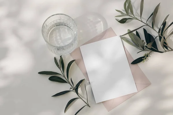 Modern summer wedding stationery still life. Drinking glass with shadows. Blank blush pink envelope, card mock up scene. White table background in sunlight. Green olive tree branches. Flat lay, top — Stock Photo, Image