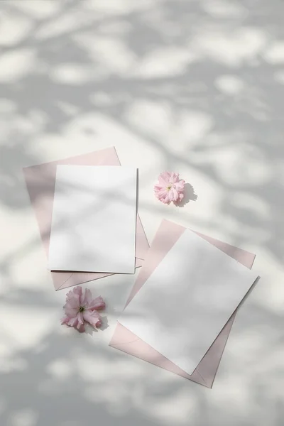 Natura morta di matrimonio. Scena femminile della colazione primaverile. Ciliegio giapponese rosa, fiori di sakura alla luce del sole. Biglietto d'auguri vuoto, invito. Buste rosa arrossite. Sfondo tavolo bianco tamponato — Foto Stock