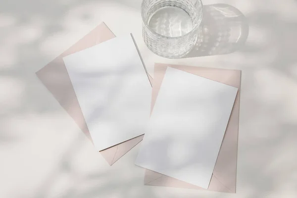 Summer wedding stationery. Blank greeting cards, invitations mockups. Blush pink envelopes and glass of water, cocktail in sunligth. Shadows overlay, white dappled table background. Flat lay, top view