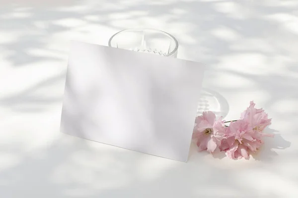 春の結婚式の文房具 日光の下でピンク色の桜と水のガラスを咲かせます 空のグリーティングカード招待状 名刺Rsvpモックアップ白いテーブル影オーバーレイ — ストック写真