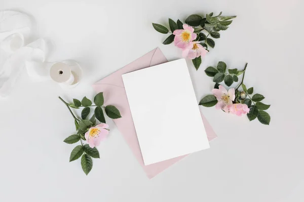 Summer wedding stationery mock-up scene with silk ribbon. Blank greeting card, invitation, pink envelope and blooming dog rose flowers isolated on white table background, flat lay, top view.
