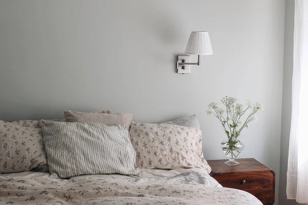 Bedroom view. Striped and floral linen pillows, blanket. Wooden night stand, bedside table Blooming bishops goutweed flowers in glass vase. Mint, sage green walll with white lamp, elegant interior.