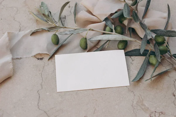 Summer branding, wedding stationery. Closeup of blank business card, invitation mock up on beige marble background. Olive tree branches and fruit with silk ribbons, top view, selective focus.