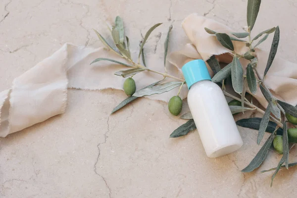 Summer cosmetic still life scene. Oil, shampoo or cream bottle with blue cap. Olive tree branch, fruit and silk ribbon on beige marble background. Moisturizer mockup, Mediterranean beauty, spa concept