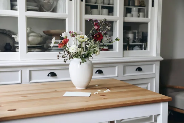 Cozy elegant Swedish Scandinavian style living room, home office. Vintage white cupboard with crockery. Wooden table. Vase of zinnia, cosmos and dahlia flowers and blank greeting card, linen sofa.