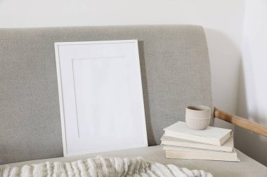 Empty wooden vertical picture frame mockup. Cup of coffee on pile of old books. Midcentury linen sofa. White wall background. Home office concept. Scandinavian interior design, artistic display.
