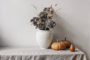 Autumn, fall still life. Big ceramic vase with dry hydrangea flowers, grass boho bouquet. Orange pumpkins and pear fruit on linen table cloth. White wall background, Halloween, Thanksgiving holiday