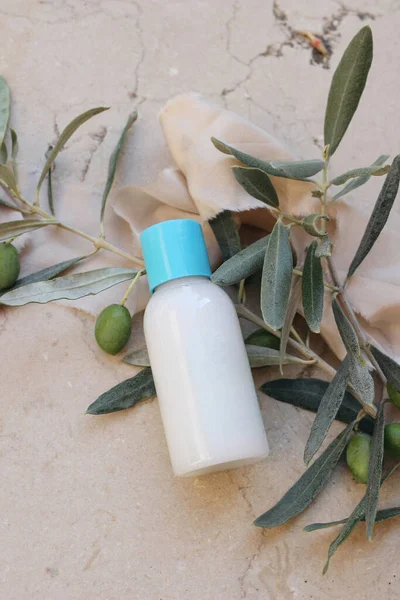 Summer cosmetic still life scene. Oil, shampoo or cream bottle mockup with blue cap. Olive tree branches, fruit on beige marble background, silk ribbon. Mediterranean beauty, spa concept, vertical top