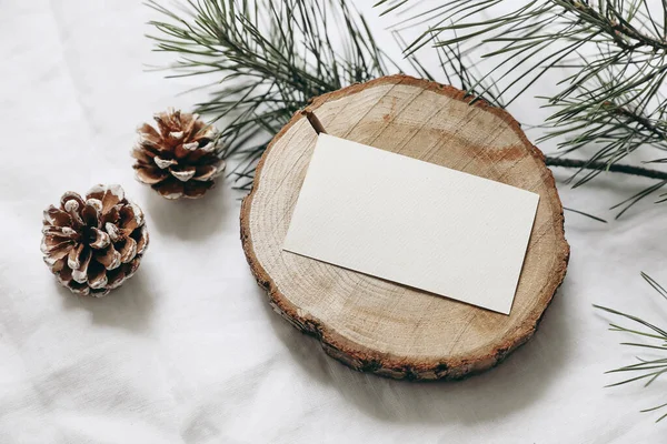 Christmas stationery still life. Blank business card, invitation mockup on cut wooden round board. Pine cones and green Christmas tree branches on white linen cloth background, top view, no people.