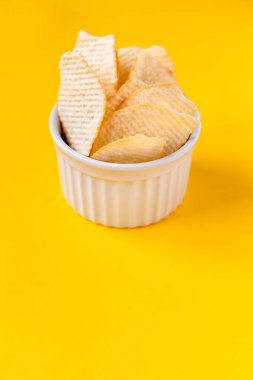 Riffled potato chips in white ceramic bowl on yellow background. Junk unhealthy food, chips, party food, fast food concept, free space for text