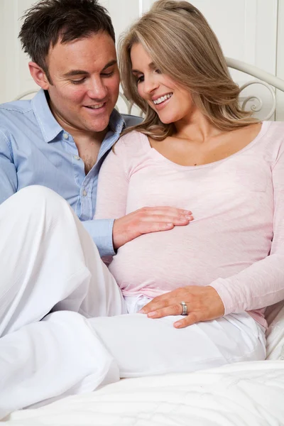 Mulher grávida feliz e parceiro sentado na cama com as mãos tocando barriga — Fotografia de Stock