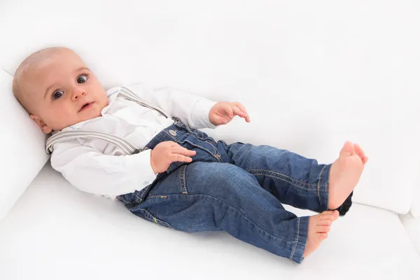 Lindo bebé acostado descalzo en un sofá blanco con vaqueros azules —  Fotos de Stock