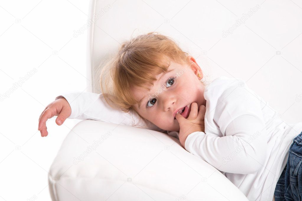 Cute child - shy girl lying on white sofa sucking thumb or finger