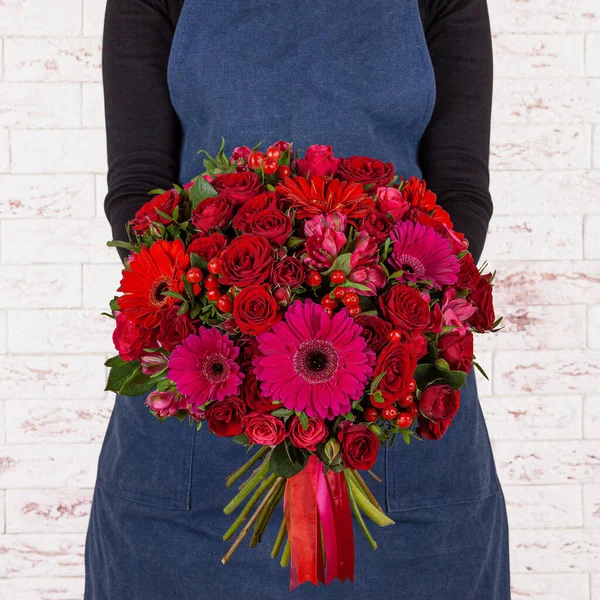 年轻的女花匠 手里拿着一束美丽的红花 上面挂着红带 墙上挂着一束鲜红的花束 免版税图库照片