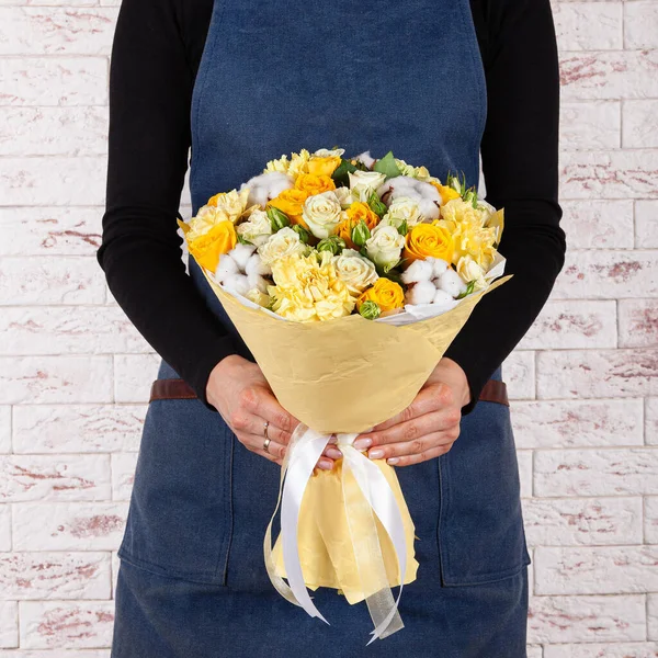 Young woman florist in blue denim apron holding bunch of flowers in paper wrap cone over brick wall background — Stockfoto