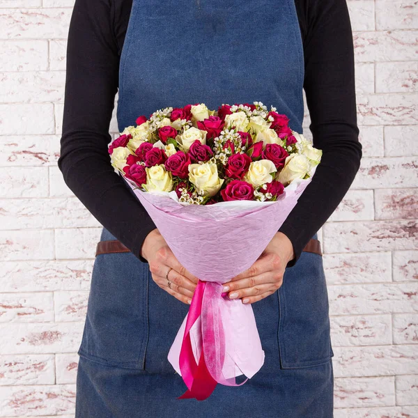 Young woman florist in blue denim apron holding bunch of flowers in paper wrap cone over brick wall background — Fotografia de Stock