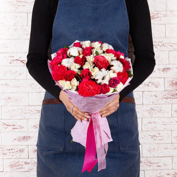 Young woman florist in blue denim apron holding bunch of flowers in paper wrap cone over brick wall background — Fotografia de Stock