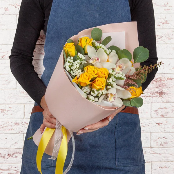 Young florist woman in blue denim apron holding bunch of yellow and white flowers bouquet over light brick wall background — Stockfoto