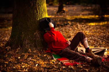 Siyah şapkalı, kırmızı kazaklı, sakallı, vahşi bir genç adam. Öğleden sonra parkta, bir sonbahar ağacının altında.