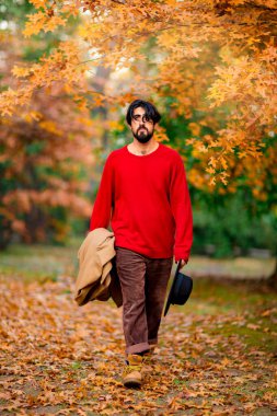 Genç bir adam, koyu saçlı, sakallı bir şapka takıyor, sonbahar güneşli bir günde parkta yürüyor.