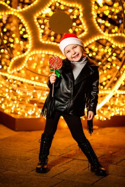 Noel Baba şapkalı güzel, neşeli küçük bir kız. Işıkların yanında duruyor. Şenlikli bir akşamda, sokakta.