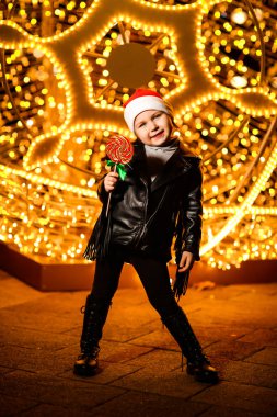 Noel Baba şapkalı güzel, neşeli küçük bir kız. Işıkların yanında duruyor. Şenlikli bir akşamda, sokakta.