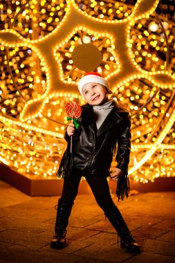 Noel Baba şapkalı güzel, neşeli küçük bir kız. Işıkların yanında duruyor. Şenlikli bir akşamda, sokakta.