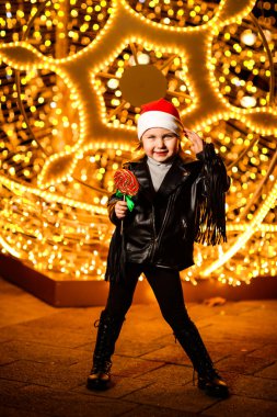 Noel Baba şapkalı güzel, neşeli küçük bir kız. Işıkların yanında duruyor. Şenlikli bir akşamda, sokakta.