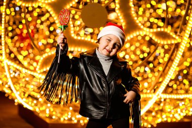 Noel Baba şapkalı güzel, neşeli küçük bir kız. Işıkların yanında duruyor. Şenlikli bir akşamda, sokakta.