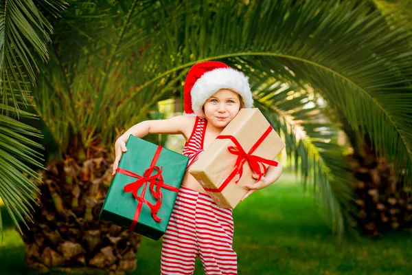 在一个温暖的国度里 一个穿着圣诞老人衣服的美丽的小女孩 手里拿着礼物 站在绿树旁过年 — 图库照片