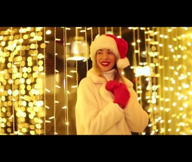 Beyaz kürklü, Noel Baba şapkalı ve kırmızı eldivenli güzel, neşeli bir kız. Parlayan ışıkların arkasında dans ediyor, gülümsüyor ve gülüyor.