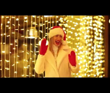 Beyaz kürklü, Noel Baba şapkalı ve kırmızı eldivenli güzel, neşeli bir kız. Parlayan ışıkların arkasında dans ediyor, gülümsüyor ve gülüyor.